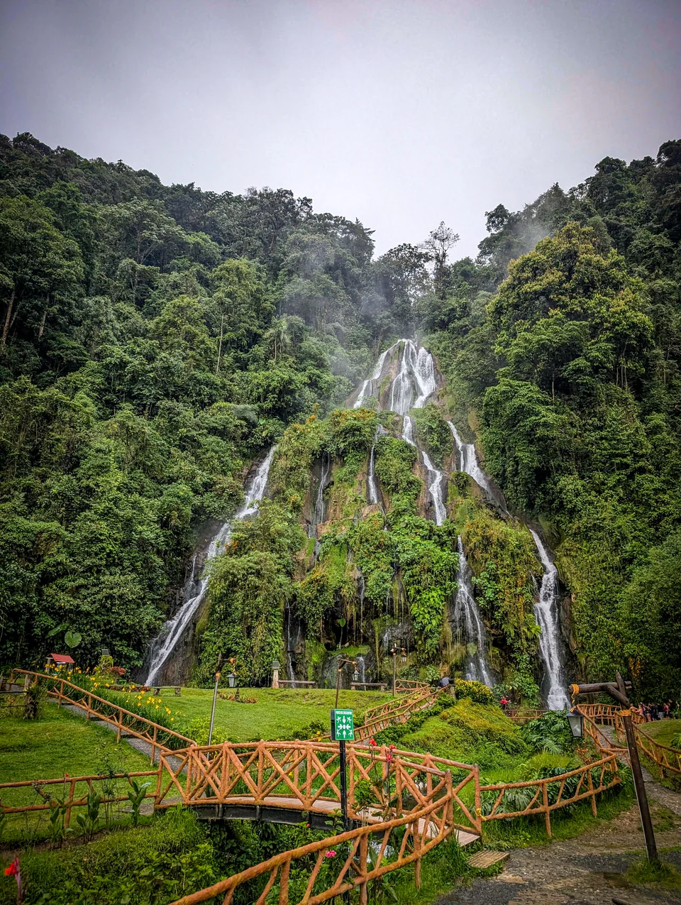 Waterfall running through dense greenery, with a wooden bridge in the foreground.
