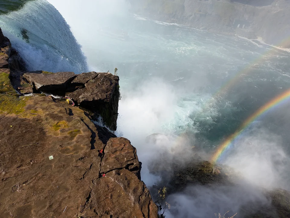 Rocky rim of a waterfall with mist and a rainbow on the right.