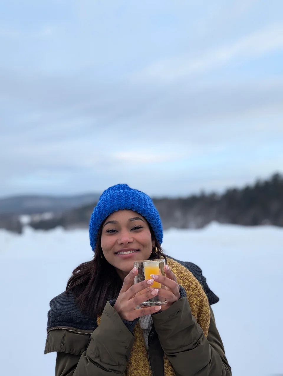 A woman in a blue hat holds an ice glass with an orange drink in front of a snowy lake.