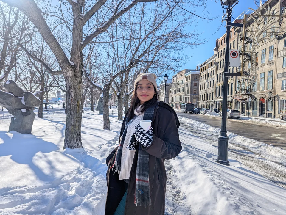 A woman in a long coat and scarf on a snowy street with stone buildings.