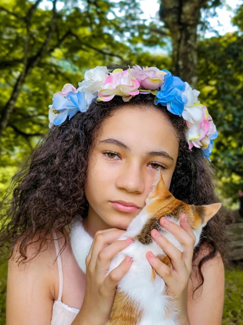 Portrait of a young woman with a flower crown holding an orange and white cat against her cheek.