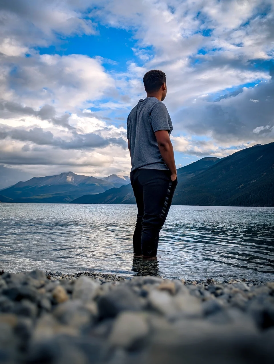 A man, seen from behind, stands with his feet in a lake, looking at mountains under a cloudy sky.