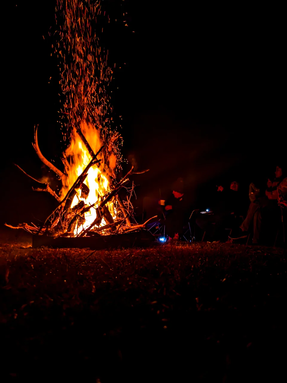 A large bonfire burns at night, with sparks rising and silhouettes around it.