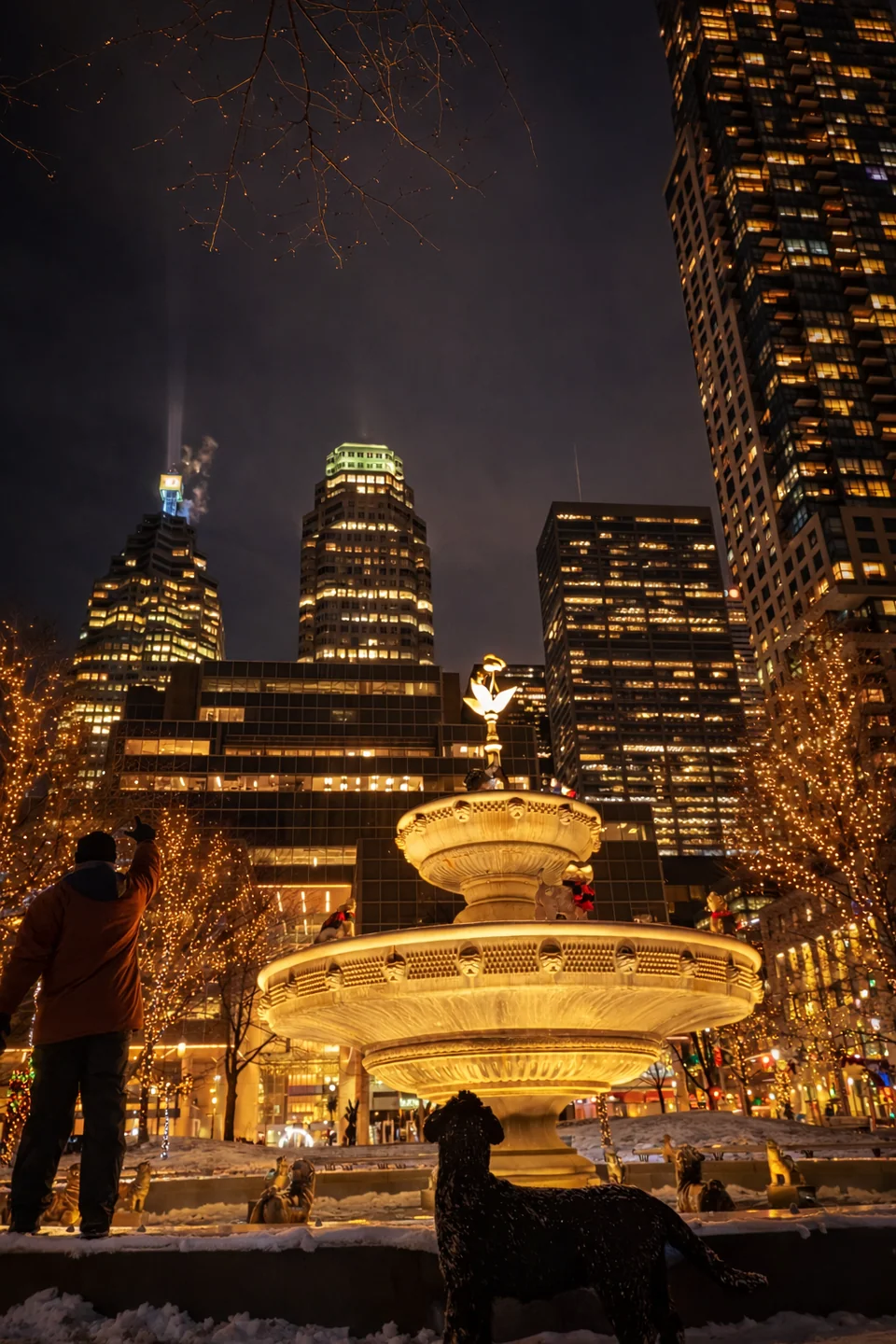 A fountain lit in gold in a night square, surrounded by skyscrapers.
