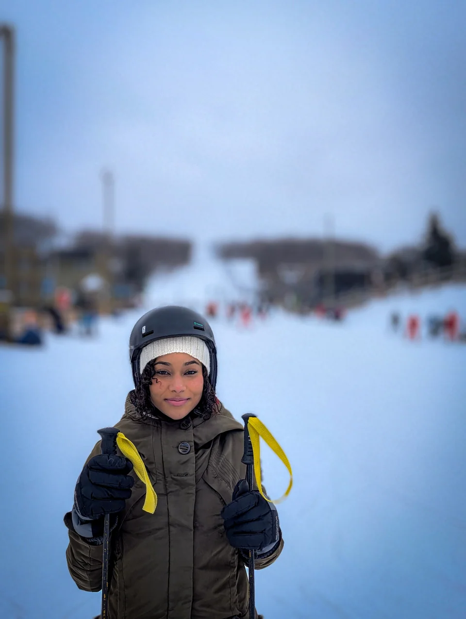 A boy in a helmet and gloves smiles on a ski slope with people behind.