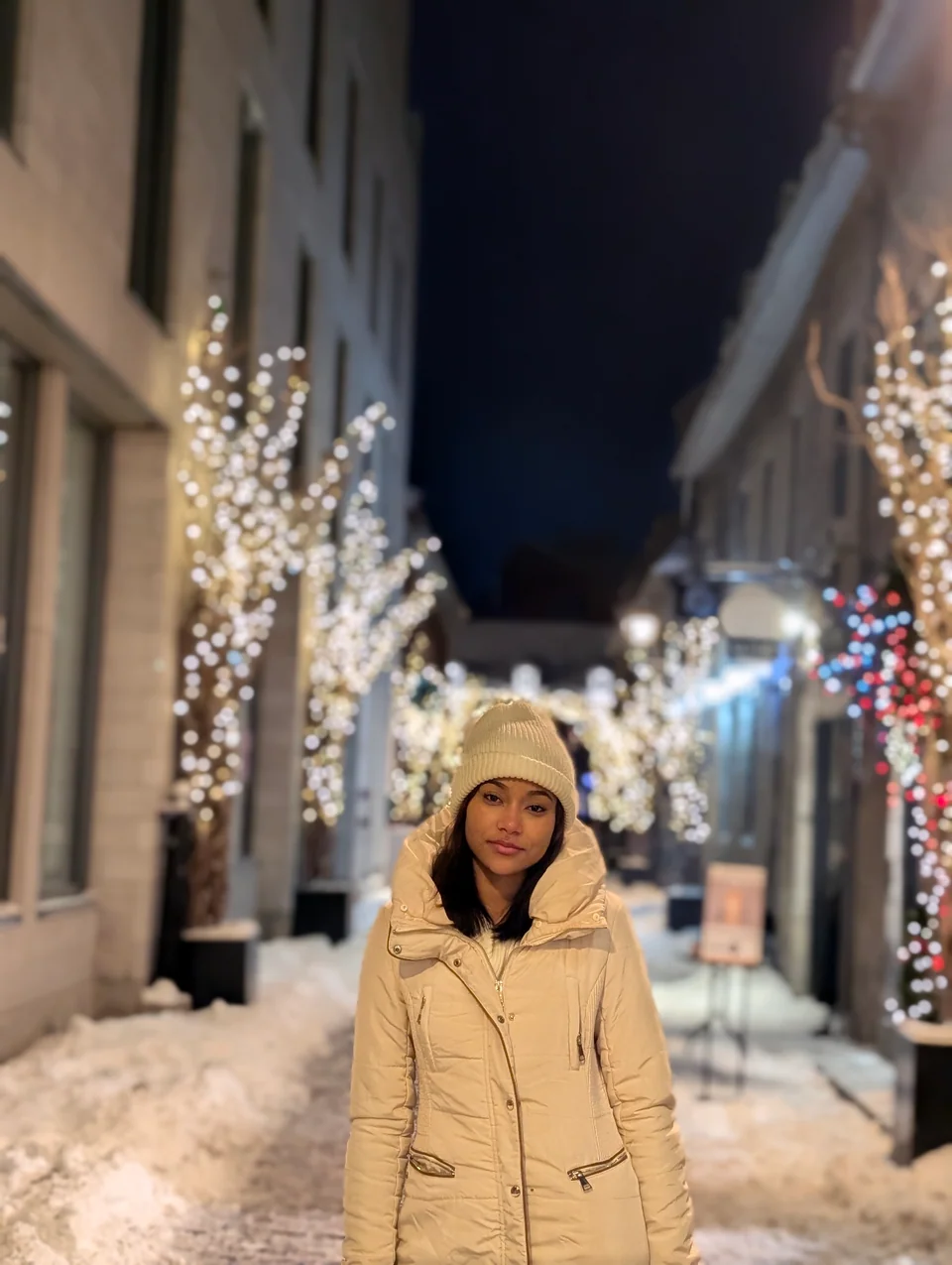 A woman in a hat and light coat on a night street with trees full of lights.