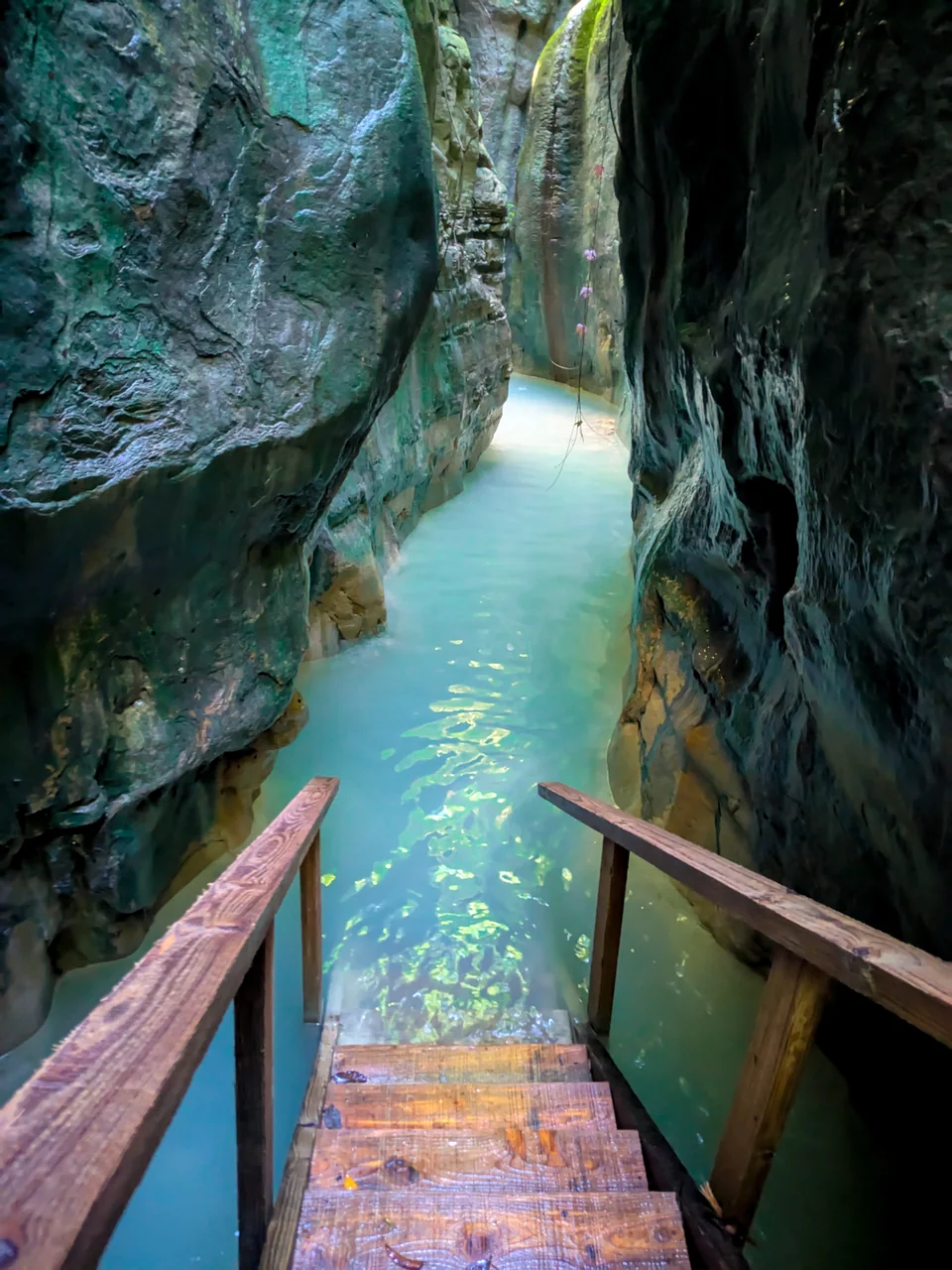 Wooden walkway over turquoise water in a narrow canyon of dark rock.