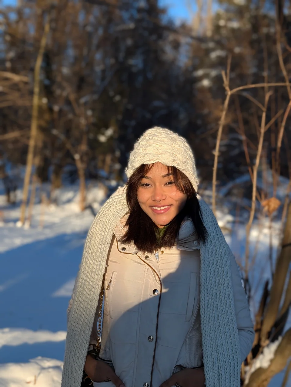 Portrait of a smiling woman in a wool hat and scarf in a sunny snowy forest.