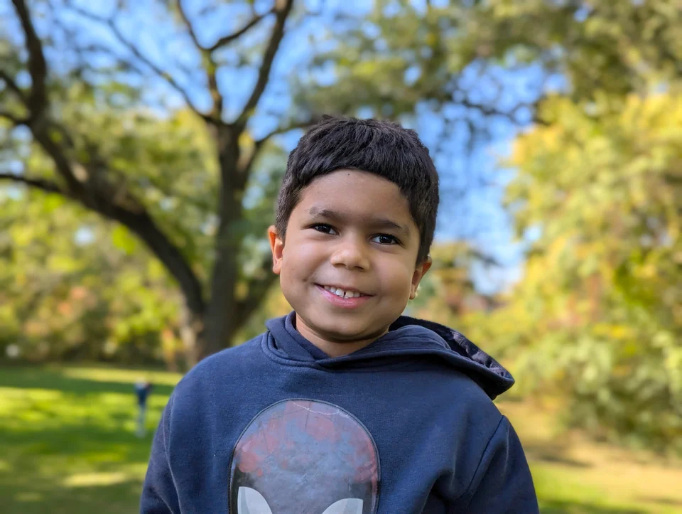 Portrait of a smiling boy in a blue sweatshirt in a park with trees.