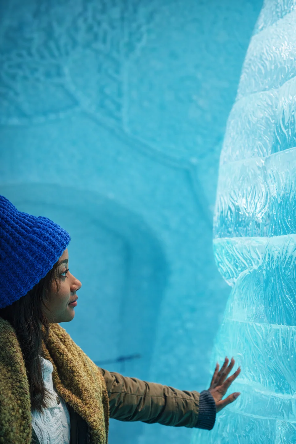 A person in a blue hat rests a hand on a carved blue ice wall.