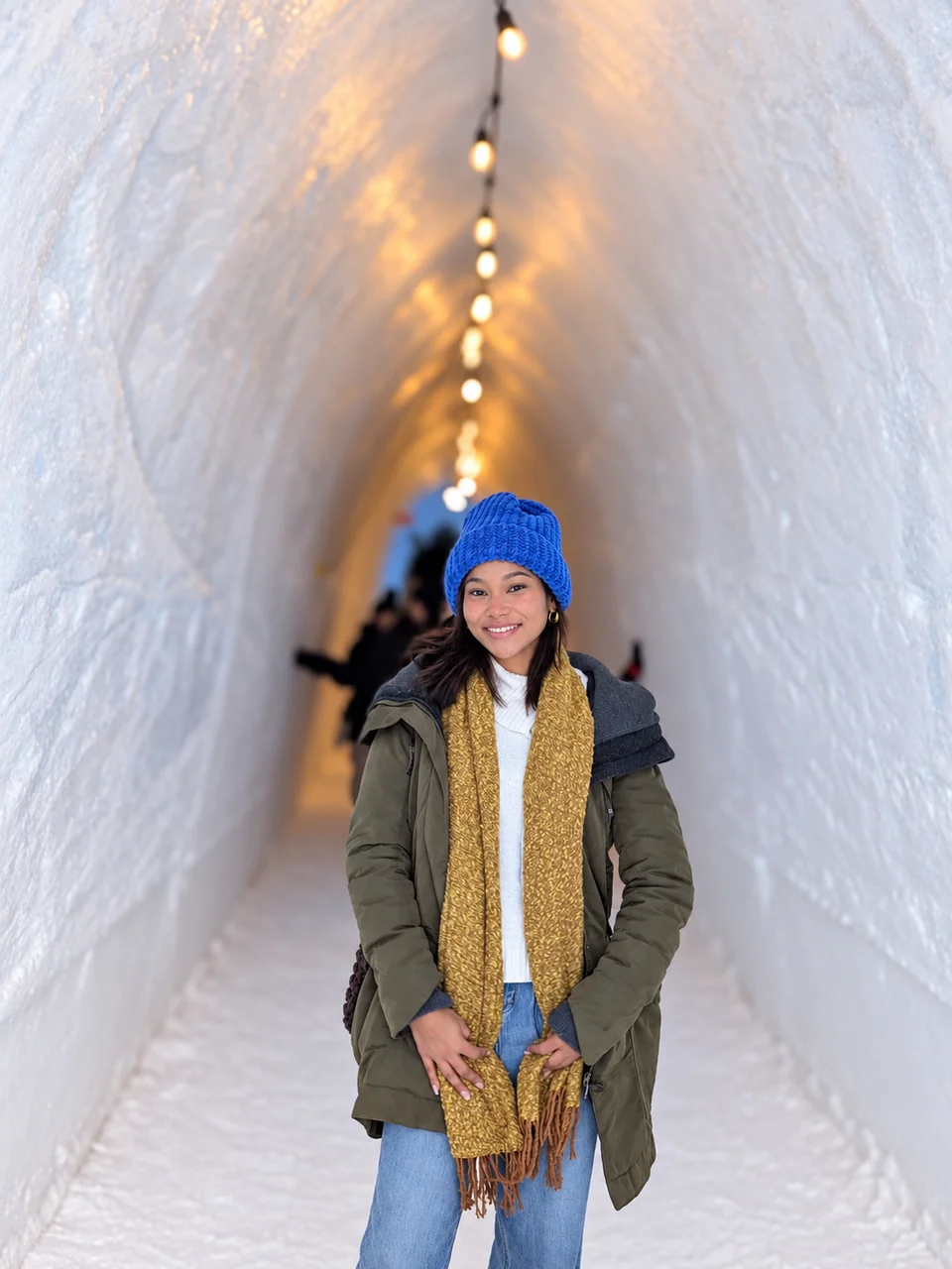 A person in a blue hat inside an ice tunnel lit by a row of warm lights.